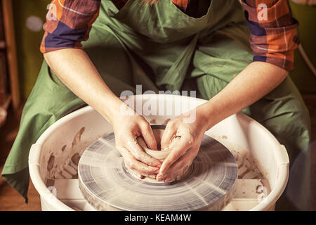 Eine Nahaufnahme eines weiblichen Potter in einem karierten Hemd und eine überdachte Schürze sitzt und formt eine Kanne braun Ton in der Werkstatt auf der Töpferscheibe, vie Stockfoto