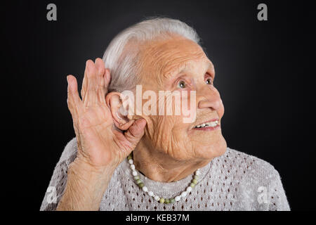 Gerne ältere Frau, die versucht, mit der Hand über Ohr über grauen Hintergrund zu hören Stockfoto