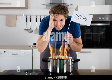 Besorgt Junger Mann Hält Hilfe Flagge Mit Utensil Auf Feuer Auf Elektroherd Zu Hause Stockfoto