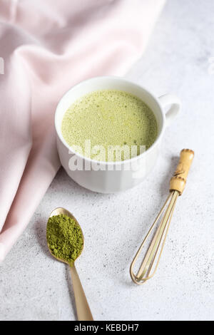 Styled Foto eines grünen Tee Matcha Latte in eine weiße Tasse mit Matcha Pulver auf einem goldenen Löffel, Bambusbesen (Chasen) und rosa Tuch Stockfoto