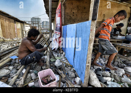 Jakarta, Java, Indonesien. August 2016. Ein Mann baut sein Haus, während sein Sohn barfuß durch die Ruinen des Pasar Ikan Viertels von Jakarta spaziert. Im April letzten Jahres stürmten Hunderte von Soldaten und Polizisten Pasar Ikan, ein historisches Fischerviertel nördlich von Jakarta, und machten alles in Trümmer. Wo es Backsteinhäuser gab, jetzt gibt es Berge von Schutt und Zelten. Die Einwohner zögern, die Nachbarschaft zu verlassen und weigern sich, in die von den Behörden gewährten Schutzhütten zu ziehen, weil sie behaupten, sie könnten sich diese nicht leisten. Das sind Vertreibungen Stockfoto