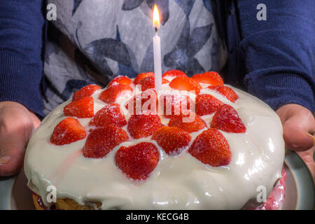 Woman's Hände halten ein Schwamm Erdbeere Geburtstag Kuchen mit Sahne und Erdbeeren auf die Oberseite, plus eine brennende Kerze. England, UK. Stockfoto