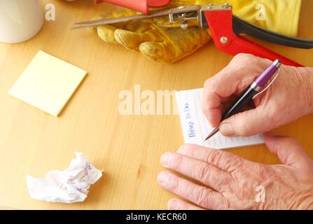 Die Gemeinkosten der Mann schreiben Sie eine Aufgabenliste mit zerknittertem Papier, Notepad, Kaffeetasse, Gartenbau Handschuh und Grass Cutter Stockfoto