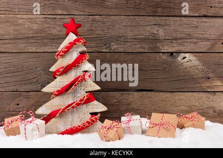 Shabby Chic Holz Weihnachtsbaum mit rustikalen Red Garland und Geschenke im Schnee gegen eine Holz Hintergrund Stockfoto