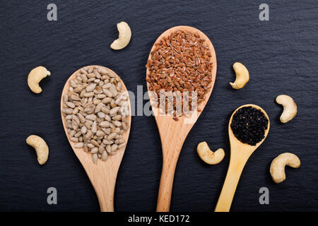 Biologische Sonnenblumenkerne, Leinsamen und Sesam in Holzlöffel und rohe Cashewnüsse auf schwarzem Schieferstein-Hintergrund mit Kopierfläche Stockfoto