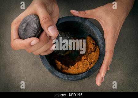 Die Frauen halten mit Mörser und Pistill und Spice Red Curry Paste Wirkstoff von Thai beliebte Speisen auf dunklen konkreten Hintergrund. Stockfoto