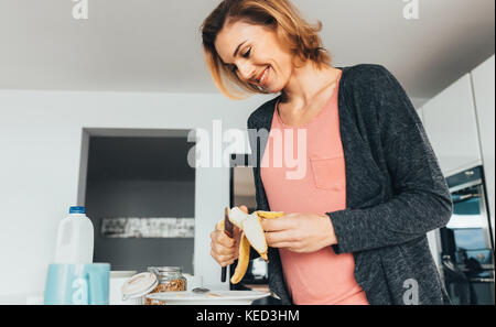 Frau vorbereiten ein gesundes Frühstück in Ihrer Küche. Lächelnde Frau hinzufügen Bananenscheiben zu ihrem Teller mit Haferflocken. Stockfoto