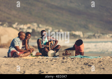 Afro-amerikanische junge Mann mit Gitarre singen ein Lied für seine Freunde. Eine Gruppe von Freunden auf Urlaub am Strand. Stockfoto