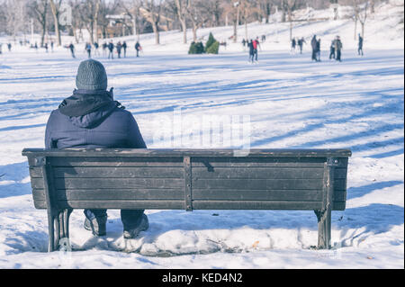 Mann sitzt auf der Bank mit Blick auf Leute Eislaufen Stockfoto