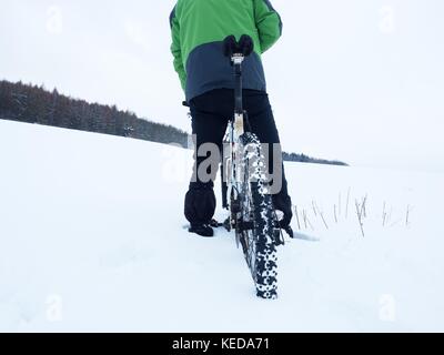 Mann mit Mountainbike Aufenthalt im Pulverschnee. verloren weg in tiefe Schneeverwehungen. Hinterrad detail. Schneeflocken schmelzen in dunklen off road Reifen. Winter Wetter können Sie Stockfoto