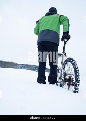 Mann mit Mountainbike Aufenthalt im Pulverschnee. verloren weg in tiefe Schneeverwehungen. Hinterrad detail. Schneeflocken schmelzen in dunklen off road Reifen. Winter Wetter können Sie Stockfoto