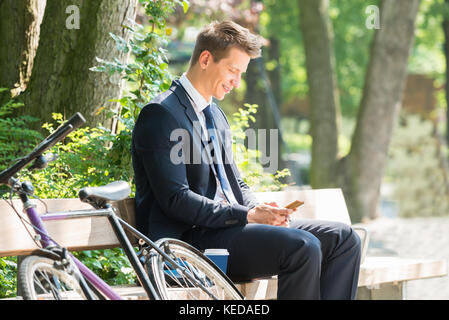 Junge männliche Geschäftsmann sitzt auf der Bank mit Handy im Park Stockfoto