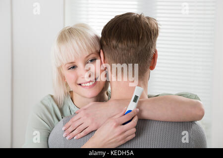 Glückliche junge Frau mit Schwangerschaft Test Mann zu Hause umarmen Stockfoto