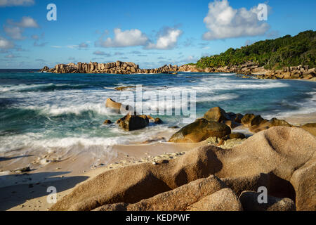 Wellen gegen golden Granitfelsen vor einem Dschungel. rauhe Wildnis - eine andere Seite der Paradies auf den Seychellen. Stockfoto