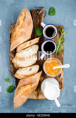 Frühstück eingestellt. Baguette, Orange Marmelade und Kaffee in Tassen auf rustikalen Holzbrett über Beton, grau-blauen Hintergrund Stockfoto