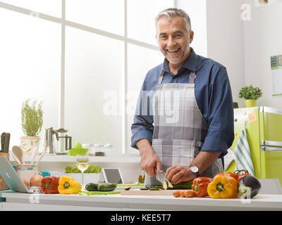 Lächelnd reifer Mann Kochen in der Küche zu Hause, er trägt eine Schürze und Schneiden frisches Gemüse, gesunde Lebensweise Konzept Stockfoto