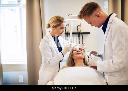 Junger Arzt und seinem Assistenten Vorbereitung einer botox Behandlung rund um die Augen einer reifen Frau lag auf einem Tisch in einer Schönheitsklinik Stockfoto