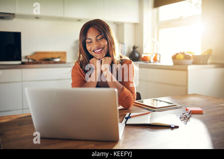 Lächelnden jungen afrikanischen weibliche Unternehmer sitzen an ihrem Küchentisch mit einem Laptop von zu Hause aus arbeiten Stockfoto
