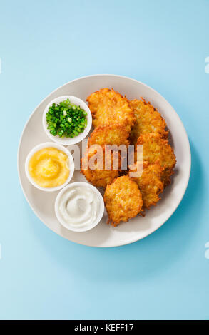 Kartoffelpuffer, latkes oder Boxty und Saucen aus Sauerrahm, Joghurt, Apfelmus und fein gehackten Frühlingszwiebeln auf einem blauen Hintergrund Stockfoto