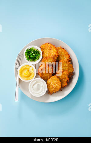 Kartoffelpuffer, latkes oder Boxty und Saucen aus Sauerrahm, Joghurt, Apfelmus und fein gehackten Frühlingszwiebeln auf einem blauen Hintergrund Stockfoto