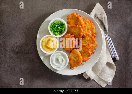 Kartoffelpuffer, latkes oder Boxty und Saucen aus Sauerrahm, Joghurt, Apfelmus und fein gehackten Frühlingszwiebeln auf grauem Stein Hintergrund Stockfoto