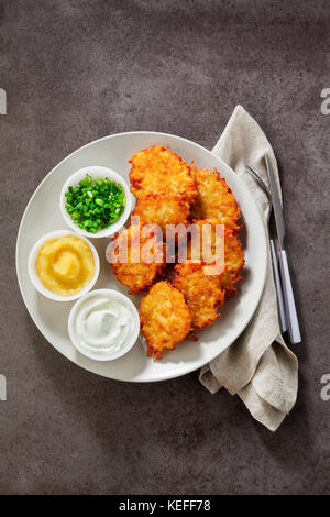 Kartoffelpuffer, latkes oder Boxty und Saucen aus Sauerrahm, Joghurt, Apfelmus und fein gehackten Frühlingszwiebeln auf grauem Stein Hintergrund Stockfoto