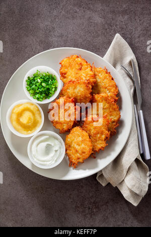 Kartoffelpuffer, latkes oder Boxty und Saucen aus Sauerrahm, Joghurt, Apfelmus und fein gehackten Frühlingszwiebeln auf grauem Stein Hintergrund Stockfoto