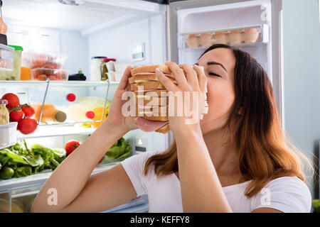 Junge Frau, Fleisch zu essen Sandwich vor dem offenen Kühlschrank Stockfoto