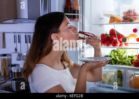 Junge Frau isst ein Stück leckeren Kuchen vor dem offenen Kühlschrank in der Küche Stockfoto