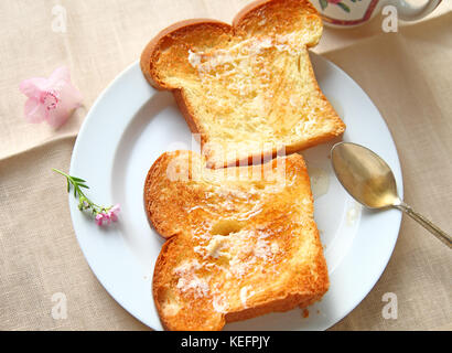 Zwei Scheiben Brot mit Butter bestrichenen Brioche mit ungefilterten Honig auf weißen Teller mit Löffel und Teetasse Stockfoto