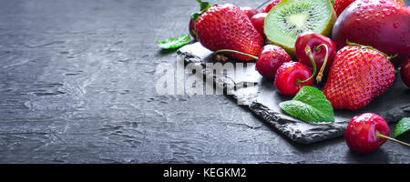 Mischung aus saftigen Sommer Früchte und Beeren. Erdbeere, Kirsche, Kiwi und Pfirsiche auf schwarzem Schiefer Platte. Lange banner Format Stockfoto