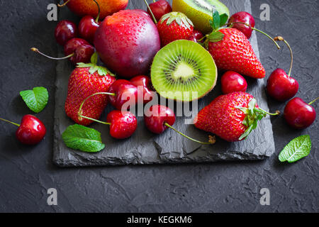 Sortiert von saftigen Sommer Früchte und Beeren. Erdbeere, Kirsche, Kiwi und Pfirsiche auf schwarzem Schiefer Platte. Stockfoto