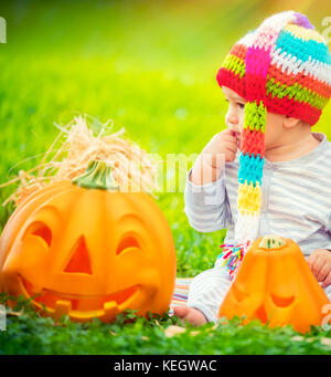 Porträt eines kleinen hübschen Kind sitzen auf der grünen Wiese in der Nähe von zwei süße Kürbisse mit geschnitzten Gesichtern, feiern Halloween Urlaub Stockfoto