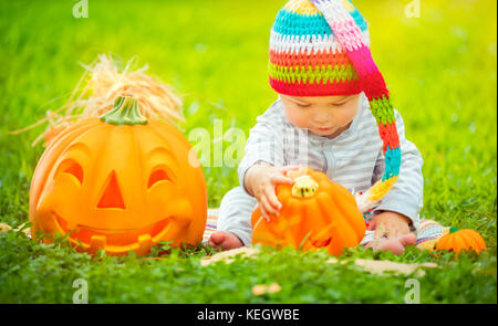 Kleines hübsches Kind sitzen auf der grünen Wiese in der Nähe von zwei süße Kürbisse mit geschnitzten Gesichtern, feiern Halloween Urlaub Stockfoto