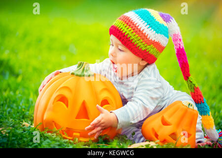 Süße kleine baby boy trug lustig bunte hat versucht zu beißen schöne geschnitzte festliche Kürbis, Spielen im Freien mit traditionellen halloween Spielzeug Stockfoto