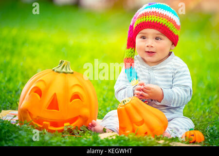 Happy Baby auf Halloween Urlaub im Freien, sitzend auf grünem Gras Hinterhof, tragen Lustig festlich bunten Hut, spielen mit niedlichen geschnitzte Kürbisse Stockfoto