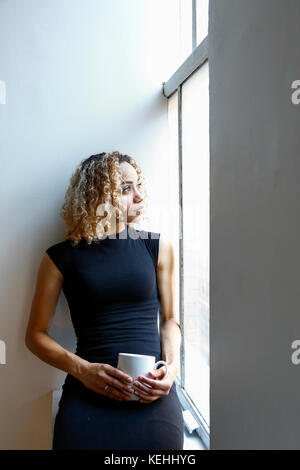 Nachdenkliche Mischrassenfrau, die auf der Fensterbank sitzt und Kaffee trinkt Stockfoto