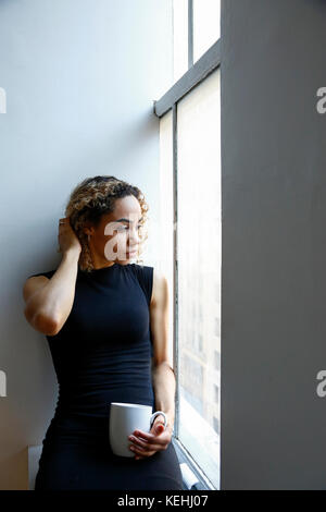 Nachdenkliche Mischrassenfrau, die auf der Fensterbank sitzt und Kaffee trinkt Stockfoto