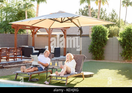 Kaukasisches Paar liest Zeitung und benutzt Laptop in der Nähe des Swimmingpools Stockfoto