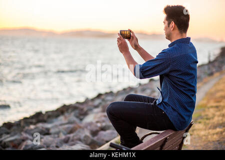 Chinesischer Mann sitzt auf der Bank fotografieren Meer mit Handy Stockfoto