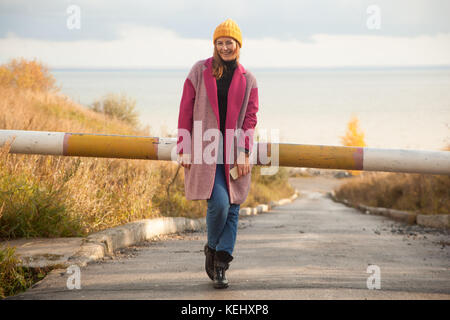 Junge schöne Brünette Frau in Gelb strick Stirnband, rosa Jacke und Jeans Lächeln und posieren im Freien auf einen Tag fallen beim Gehen Stockfoto