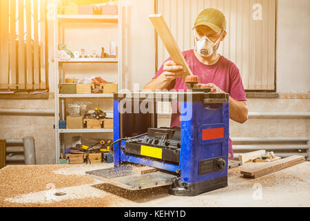 Eine männliche Tischler Builder in Arbeitskleidung, eine Maske und eine Kappe bereitet ein Brett für das Scheuern auf einem dickenhobeln Maschine in einem Licht workshop Stockfoto