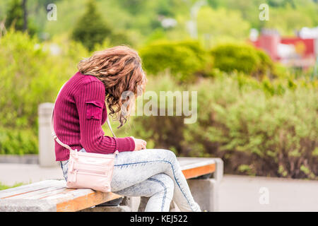 Eine junge Frau sitzt auf der Bank am Bürgersteig in grün Downtown City Park in Saguenay, Kanada, Quebec im Sommer Blick auf Telefon oder Lesen b Stockfoto