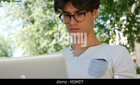 Hübscher junger Mann sitzt auf einer Holzbank im Freien arbeiten in einem städtischen Park eingeben von Informationen auf seinem Laptop. Stockfoto