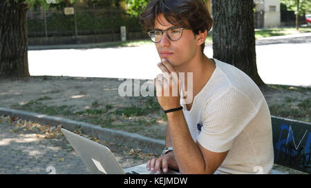 Hübscher junger Mann sitzt auf einer Holzbank im Freien arbeiten in einem städtischen Park eingeben von Informationen auf seinem Laptop. Stockfoto