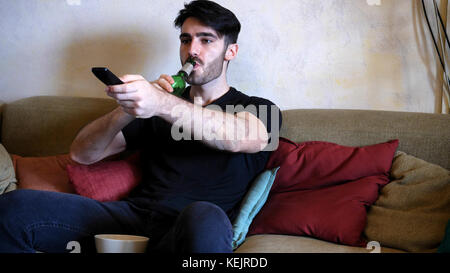 Jungen attraktiven Mann sitzt zu Hause auf der Couch im Wohnzimmer, Bier trinken und fernsehen, TV mit Fernbedienung, entspannen in einem faulen Stockfoto