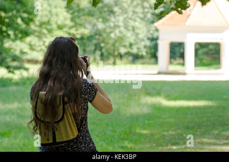 Eine Ansicht der Rückseite eine junge Frau mit Sonnenbrille und Rucksack fotografieren ein altes Gebäude in die Natur mit einem unscharfen Hintergrund Stockfoto