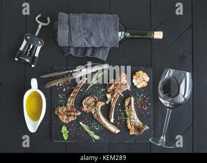 Gegrillte Lammkoteletts. Lammrücken mit Knoblauch, Rosmarin und Gewürzen auf schiefer Fach, Wein Glas, Öl in einer Untertasse, Kork screwer und Flasche über schwarzem Holz Stockfoto