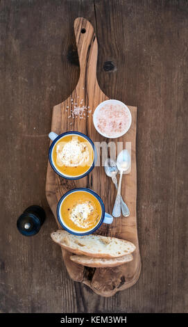 Hausgemachte Kürbis creme Suppe in Emaille Becher mit Kräutern und frischem Brot Slices auf Olivenöl, das Brett über rustikal Hintergrund, Ansicht von oben Stockfoto