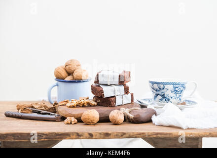Chocolate Brownie Schichten in Papier gewickelt und mit Seil müde, Porzellan, Emaille Tasse Tee Tasse Walnüsse auf rustikalen Holztisch, weißer Hintergrund Stockfoto